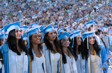 Six graduates smiling in a line, standing up.