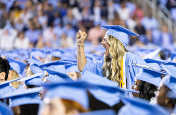 A singular student standing up, clenching their fist and yelling with excitement in a crowd of graduates.