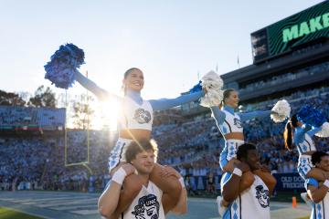 Carolina cheerleaders perform for a football stadium crowd with a sunset in the background