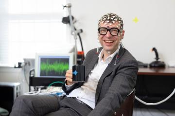 Scientist poses for photo while drinking coffee and wearing mesh device on head with white background with scientific equipment