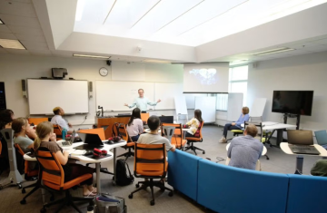 Students sit in on a class lecture with instructor standing at the front of the room