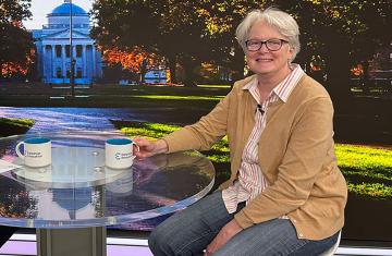 Barbara Vilen sits at a glass table with mug and campus scenery in the background