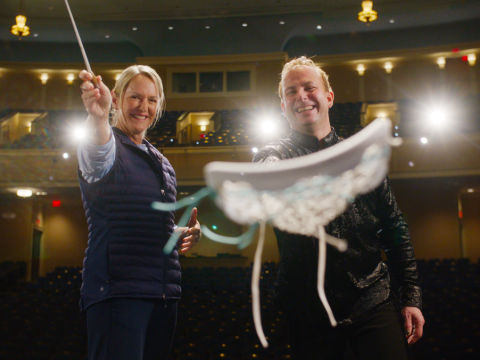 Two people stand on a theater stage smiling toward the camera, one holding a conductor&rsquo;s baton and the other presenting a decorated cap in the foreground.