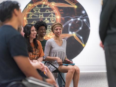 Three students smile during a group discussion in front of a large circular mural.
