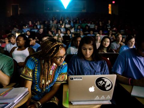 Students look at a laptop screen in a lecture classroom