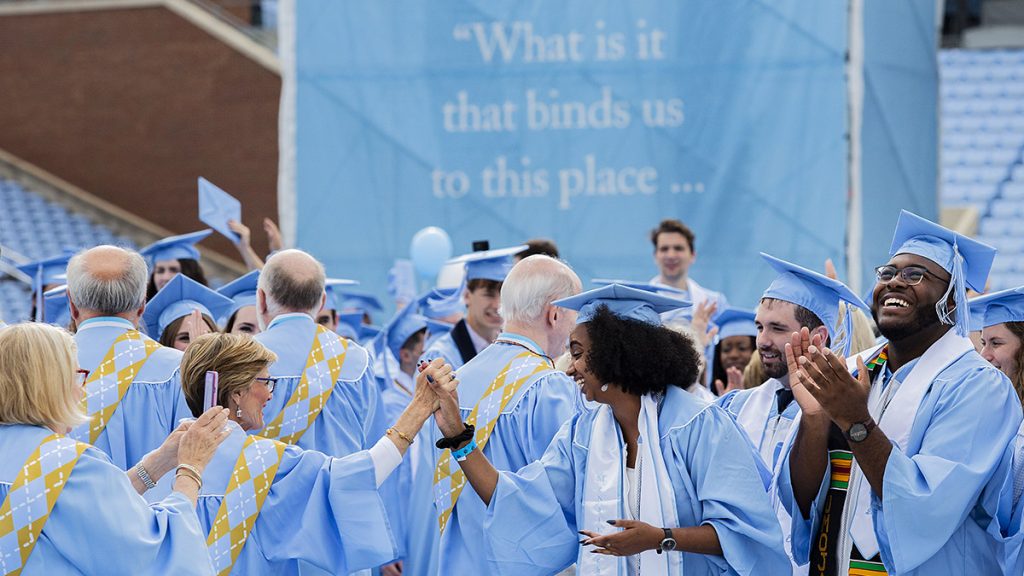 A diverse crowd of Carolina graduates, in caps and gowns, clap and shake hands at a Carolina commencement ceremony
