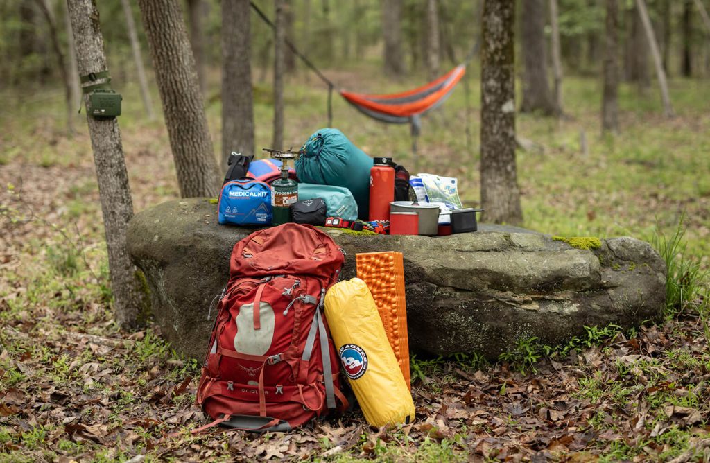 Outdoor camping equipment is arranged artfully on a large rock in the woods with a hammock in the background